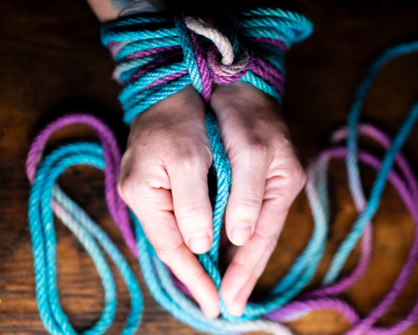 Hands bound in turquoise and violet multicolor rope, loose coils on a wooden table